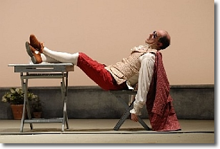 un attore sul palcoscenico di un teatro è appoggiato su una piccola sedia e tiene i piedi su di un tavolino